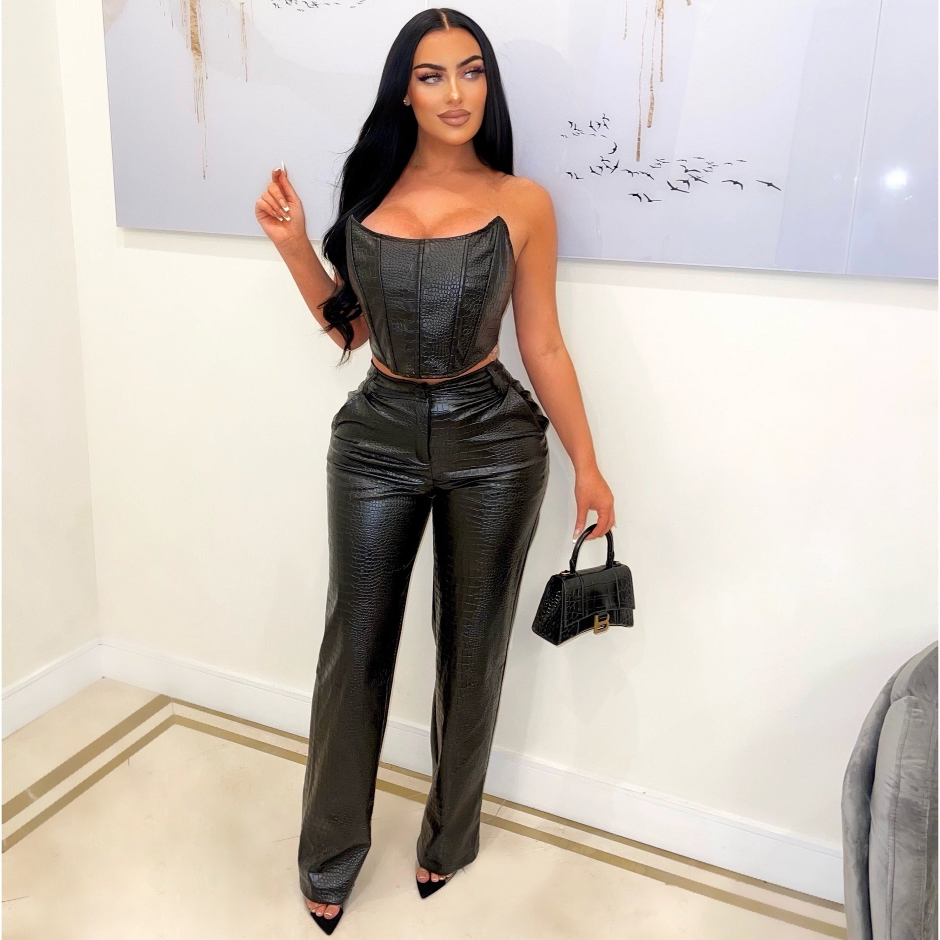 Woman in black leather outfit holding a handbag against a white wall.